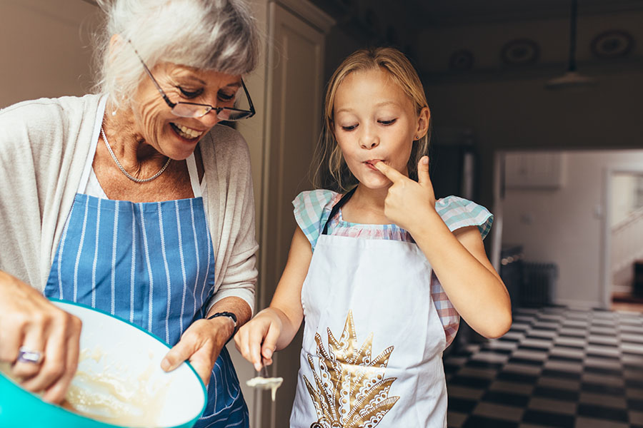 About Our Agency - View of Smiling Grandmother and Granddaughter Standing in the Kitchen Baking Together