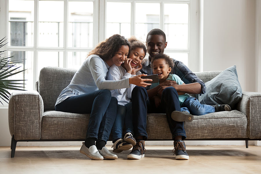 Client Center - Portrait of a Cheerful Family with Two Kids Sitting in the Living Room Having Fun Watching Videos on a Phone
